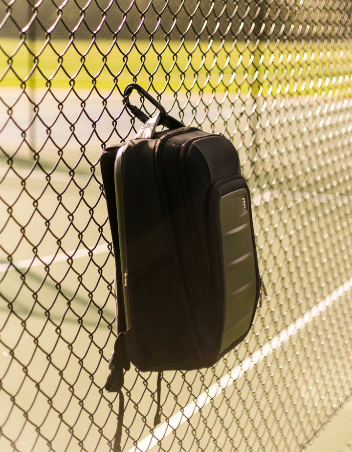 Ethos Pickleball backpack hanging on a chain-link fence with a tennis court in the background